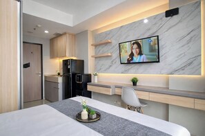 A modern hotel room with a flat-screen TV mounted on a marble wall, a dining area with a table and chairs, a kitchen with a refrigerator and microwave, and a wooden door. - Wooden Accents Studio At Monroe Tower Apartment (Cikarang)
