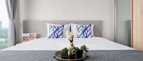 A modern bedroom with a large bed, two pillows with blue abstract patterns, a bedside table with a book and a vase with flowers, and a view of greenery outside.