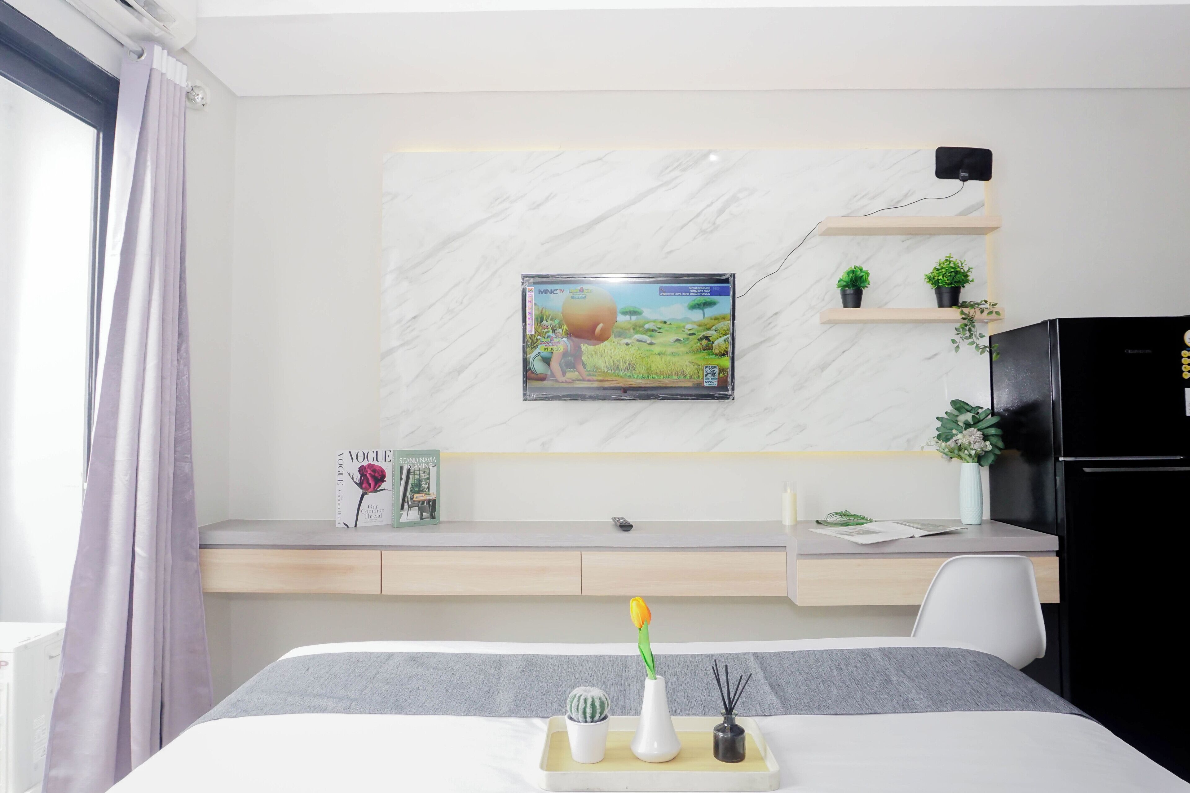 A hotel room with a bed, a desk, a TV mounted on the wall, a small shelf with plants, and a refrigerator.