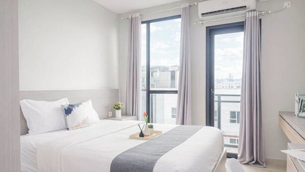 Ein modernes Hotelzimmer mit einem großen Bett, einem Nachttisch mit Blumenvase und Blick auf die Stadt durch große Fenster.