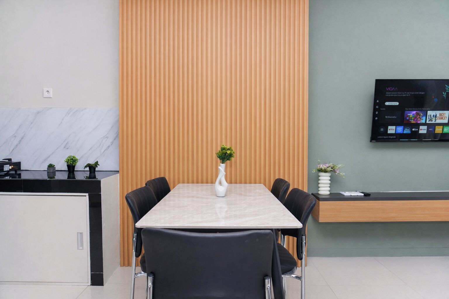A modern meeting room with a marble table, black chairs, a wooden panel wall, a mounted TV, and small potted plants.