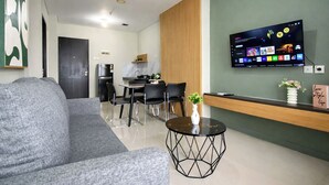 A modern living room with a grey sofa, a dining area with a table and chairs, a kitchen in the background, and a flat-screen TV mounted on the wall. - Tranquil 2Br For 6 Pax At Nifarro Park Apartment (Jakarta)
