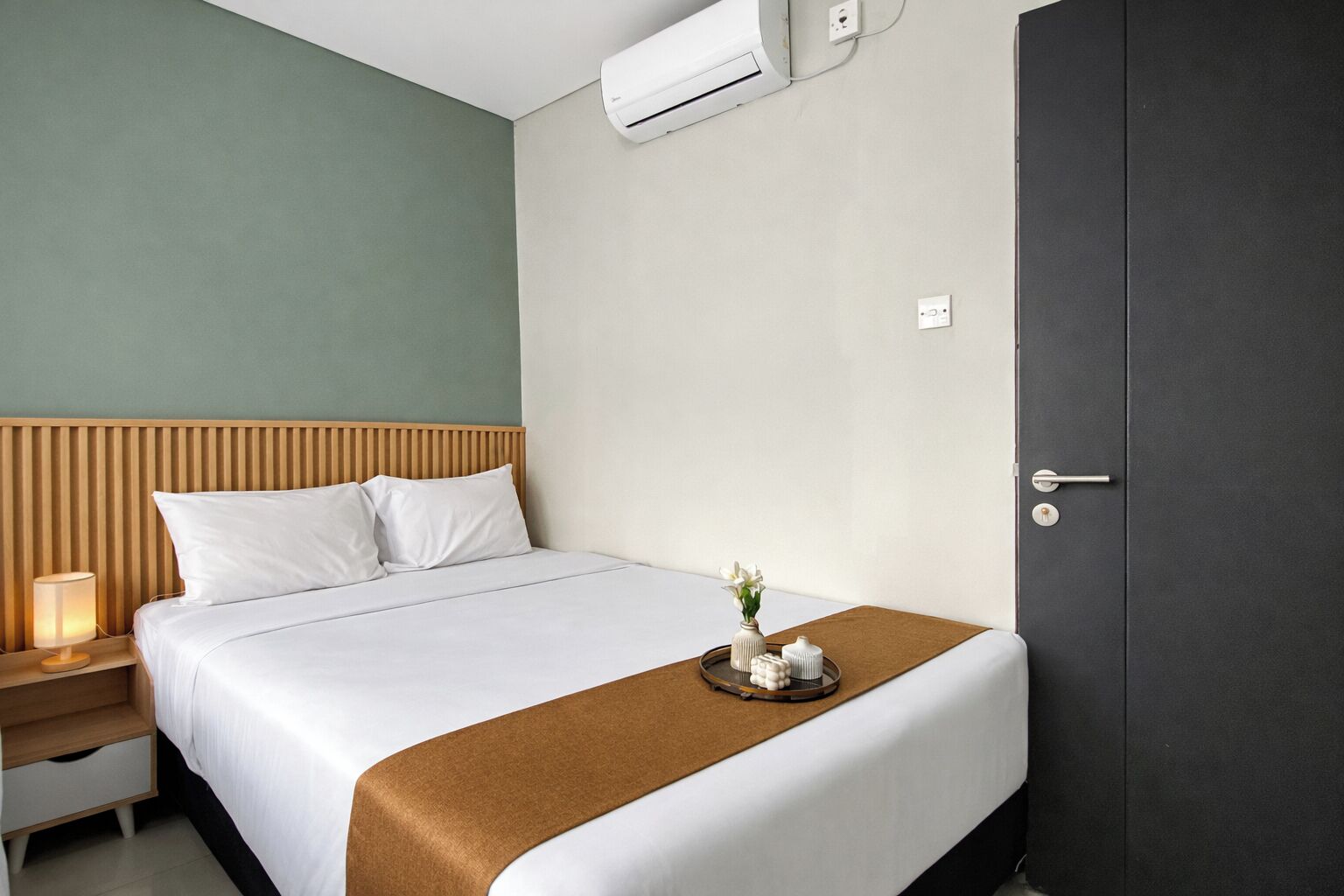 Een hotelkamer met een bed, nachtkastje, lamp en een deur.
