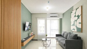 A modern living room with a grey sofa, a TV mounted on the wall, a small round table, and a window with curtains. - Tranquil 2Br For 6 Pax At Nifarro Park Apartment (Jakarta)