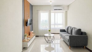 Een moderne woonkamer met een grijze bank, een ronde salontafel, een aan een houten paneel bevestigde televisie, en een stadsgezicht door grote ramen.