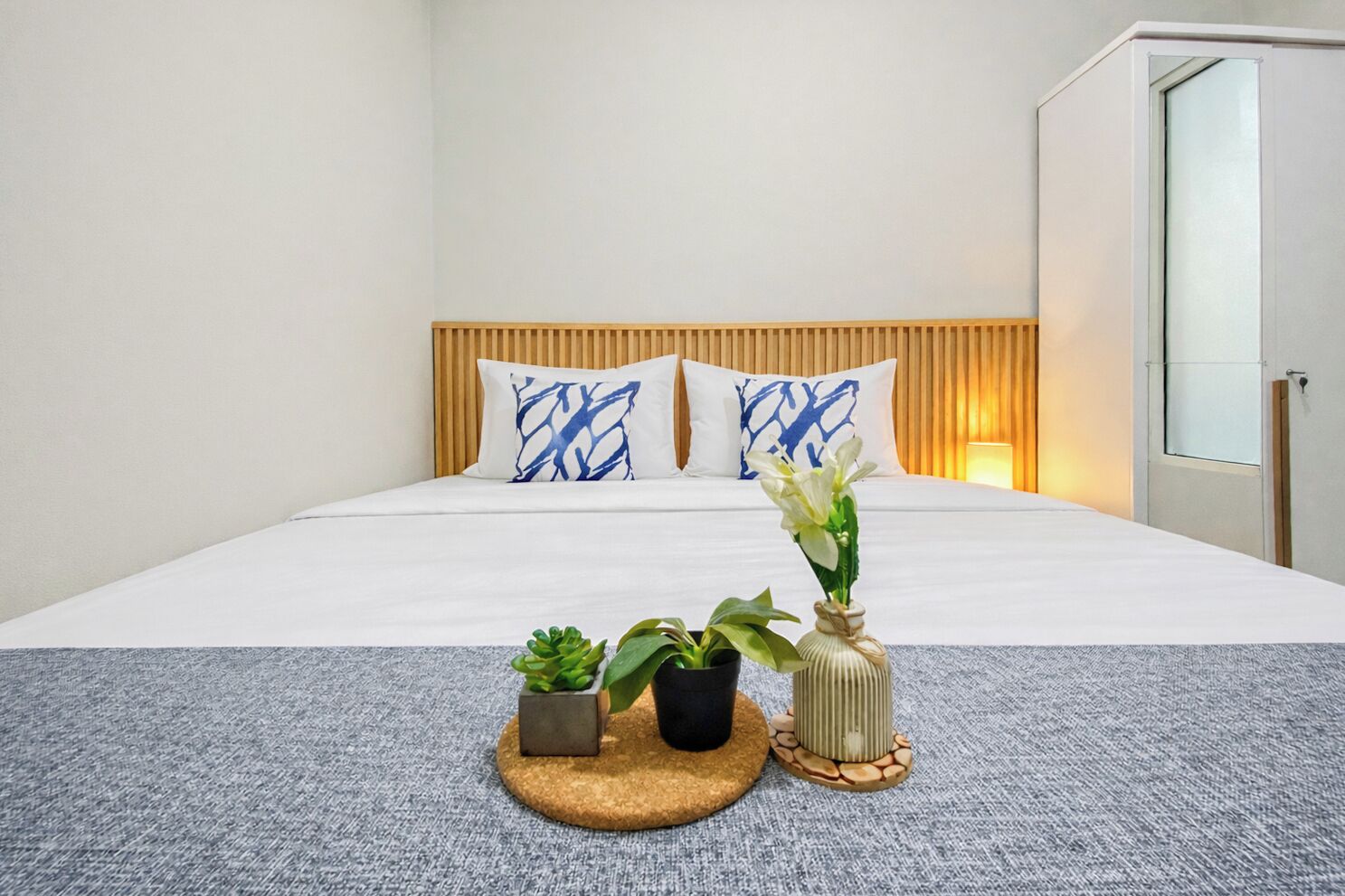 A hotel room with a bed, two pillows with blue patterns, a bedside table with a vase and plants, and a mirror on the wall.