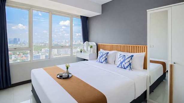 Een hotelkamer met een groot bed, een nachtkastje met een lamp, en uitzicht op de stad door grote ramen, met een deur aan de rechterkant.