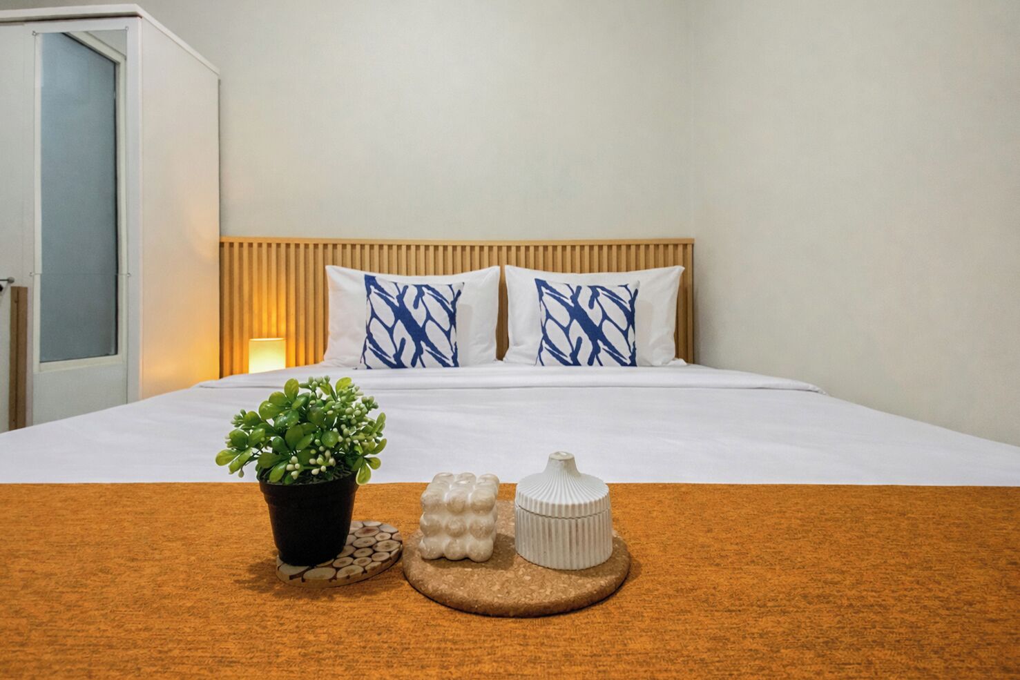 Ein Hotelzimmer mit einem Bett, zwei Kissen, einer Zimmerpflanze, einem kleinen weißen Behälter und einem hölzernen Kopfteil.