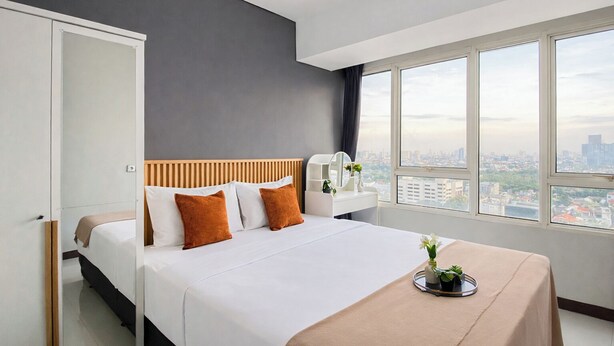 A modern hotel room with a bed, bedside table, and a view of the city through large windows. - Clean Space And Well Located 2Br At Nifarro Park Apartment (Jakarta)