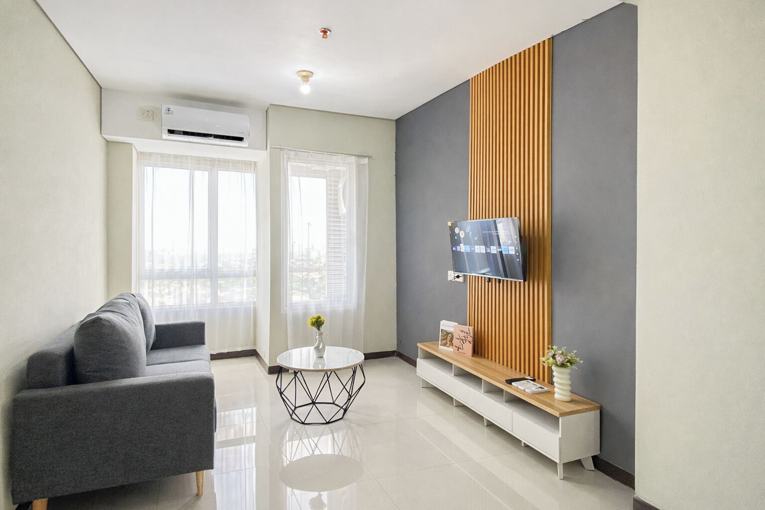 Una sala de estar moderna con un sofá, una mesa de centro redonda, un televisor montado en un panel de madera y una ventana con cortinas translúcidas.