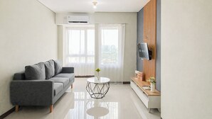 A modern living room with a grey sofa, a round coffee table, a flat-screen TV mounted on a wooden panel, and an air conditioning unit on the ceiling. - Clean Space And Well Located 2Br At Nifarro Park Apartment (Jakarta)