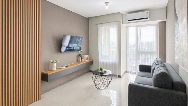 A modern living room with a flat-screen TV, a grey sofa, a wooden accent wall, and a city view through large windows. - Comfy 2Br For 6 Pax At Nifarro Park Apartment (Jakarta)