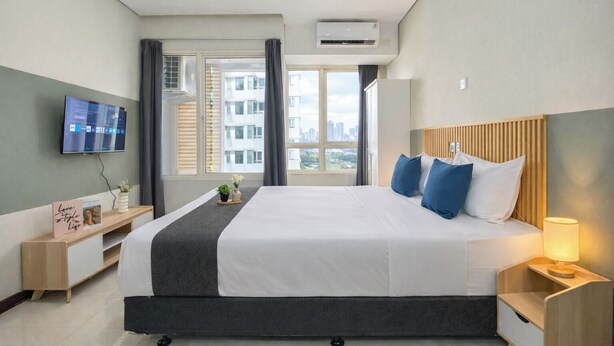 Habitación de hotel con una cama grande, un televisor en la pared, una ventana con cortinas y una mesita de noche con lámpara.