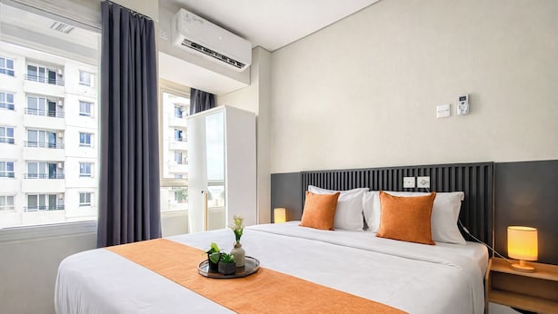 A hotel room with a large bed, a bedside table with a lamp, a window with curtains, and an air conditioning unit. - Wonderful Studio At Apartment Nifarro Park Near Pasar Minggu Station (Jakarta)