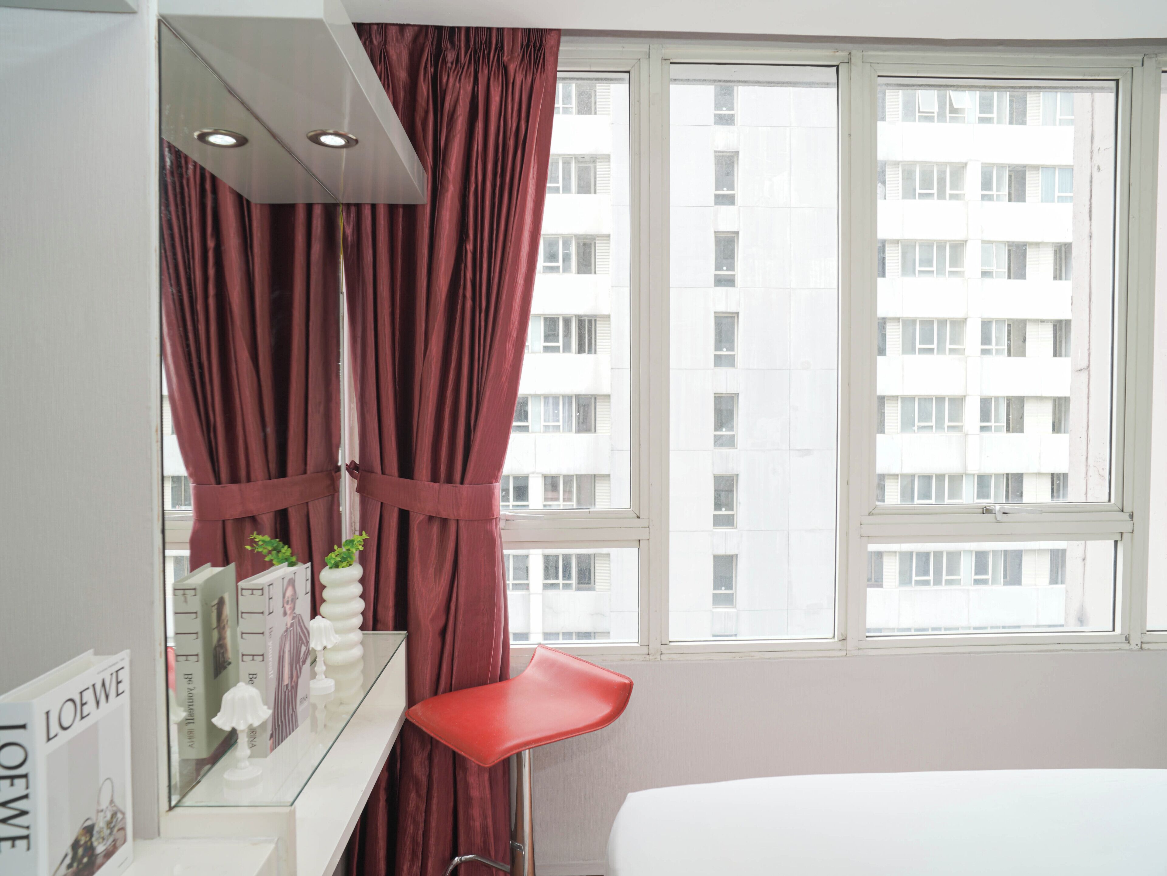 A hotel room with a bed, a red chair, a vanity with a mirror, and a window with a view of a building.