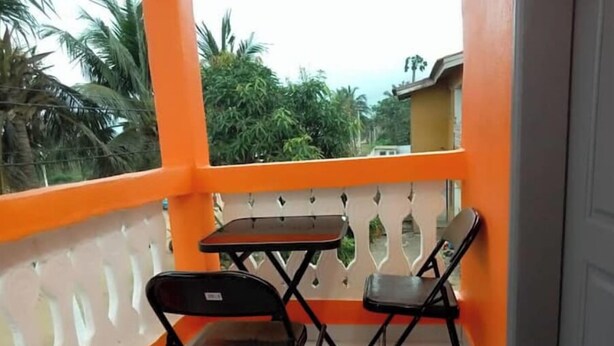 Ein Balkon mit zwei Stühlen und einem Tisch, umgeben von orangen Geländern, mit Blick auf Grünflächen und Gebäude im Hintergrund.