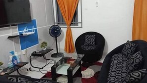 Ein Wohnzimmer mit Fernseher, Ventilator, Sessel, Tisch mit Pflanze und Fenster mit Vorhängen.
