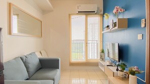 A living room with a sofa, a TV, a shelf with decorative items, and an air conditioning unit. - Cozy With Blue Accent 2Br At Podomoro Golf View Apartment (Gunung Putri)