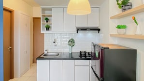 A modern kitchen with white cabinets, a black refrigerator, a hanging pendant light, and potted plants on shelves. - Cozy With Blue Accent 2Br At Podomoro Golf View Apartment (Gunung Putri)