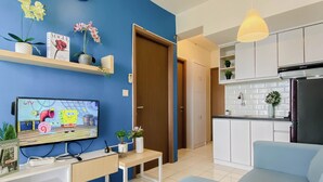 A living room with a TV, sofa, and a kitchen area with cabinets and a refrigerator. - Cozy With Blue Accent 2Br At Podomoro Golf View Apartment (Gunung Putri)