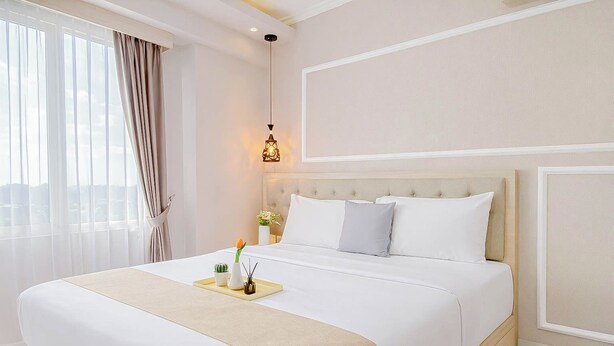 A hotel room with a large bed, a bedside table with a lamp, and a view through the window. - 2BR Corner with South and East View at Podomoro Golf View Apartment (Gunung Putri)