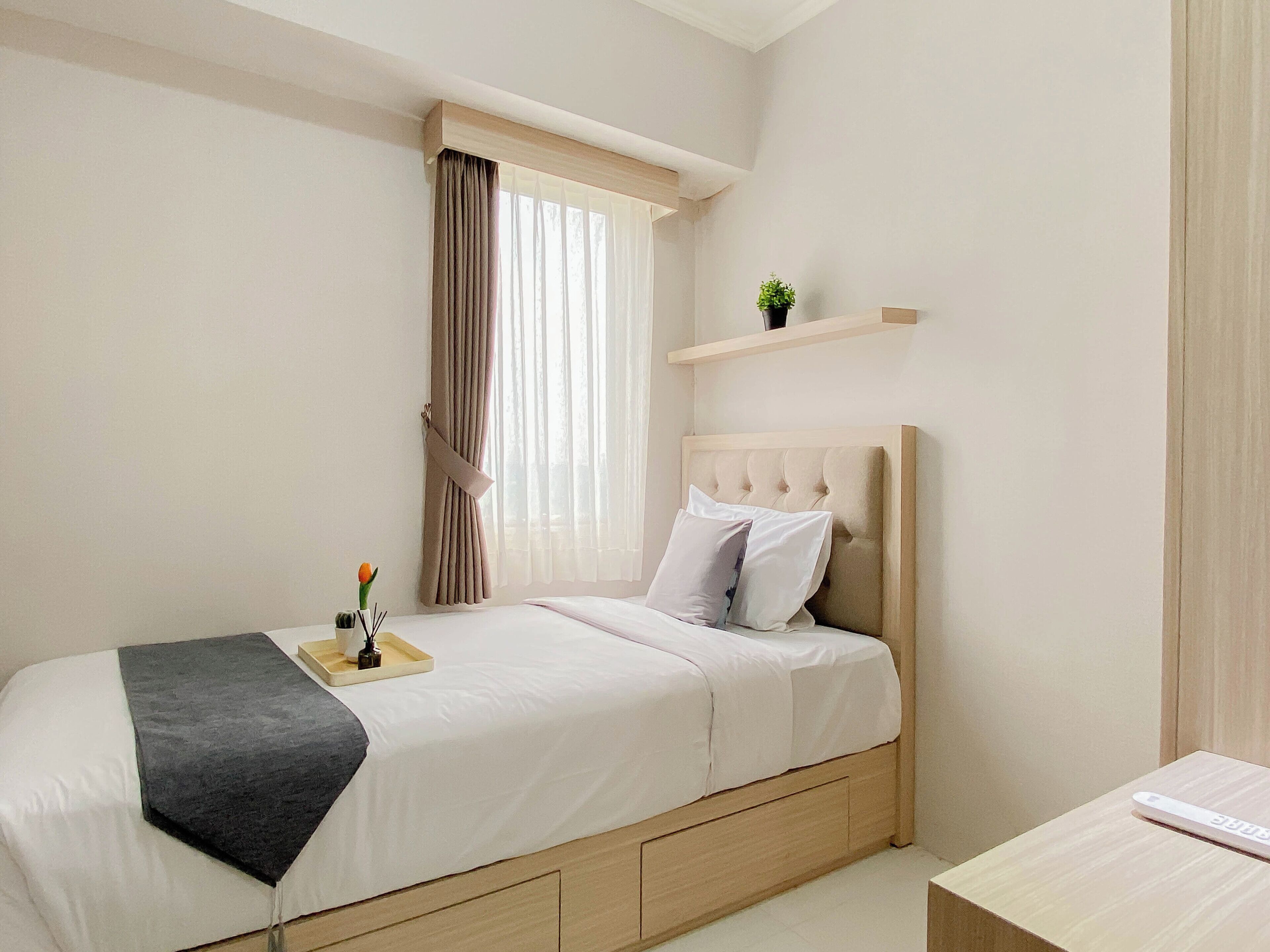 A bedroom with a bed, a bedside table, a window with curtains, and a small shelf above the bed.