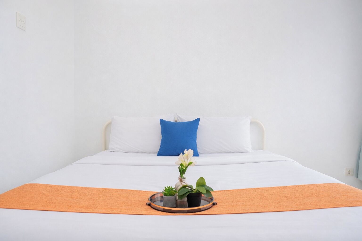 A bed with white bedding, a blue pillow, and an orange throw. There is a small potted plant on a round tray in front of the bed.