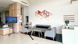 A modern lobby with a large flat-screen TV, grey sofas, a small table, and a reception desk. - Simple Style Studio At Prima Orchard Apartment (Bekasi)