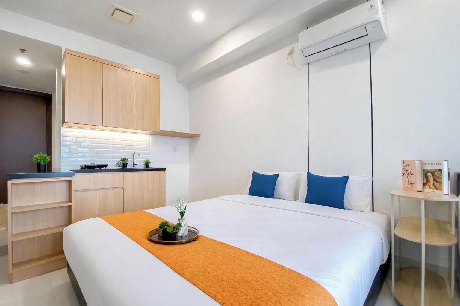A modern hotel room with a bed, a small kitchenette, and a ceiling-mounted air conditioning unit.