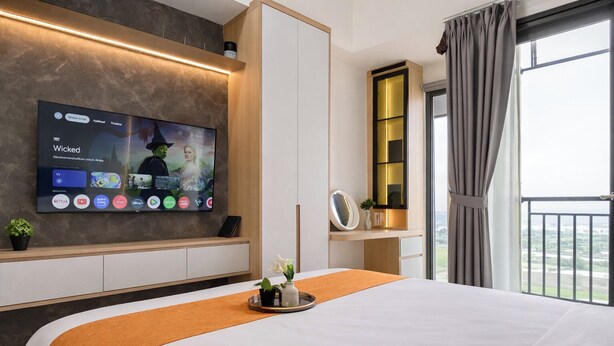 A modern hotel room with a large bed, a flat-screen TV mounted on the wall, a small potted plant, and a view of the outdoors through the window. - Modern Japandi Studio At Sayana Bekasi Apartment (Bekasi)