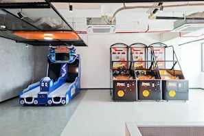 An indoor arcade with a racing game, basketball shooting game, and a claw machine. - 2Br Urban Areum Residence At Sky House Alam Sutera (Tangerang)