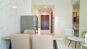 A dining area with a marble wall, a wooden table, beige chairs, a white vase with a plant, and a white chair with a cushion. - Tranquil 2Br For 3 Pax At Sky House Alam Sutera Apartment (Tangerang)