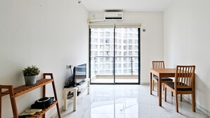 A modern living room with a TV, wooden shelves, a dining table with chairs, and a large window with a city view. - Neat 2Br For 3 Pax At Sky House Bsd Apartment (Pagedangan)