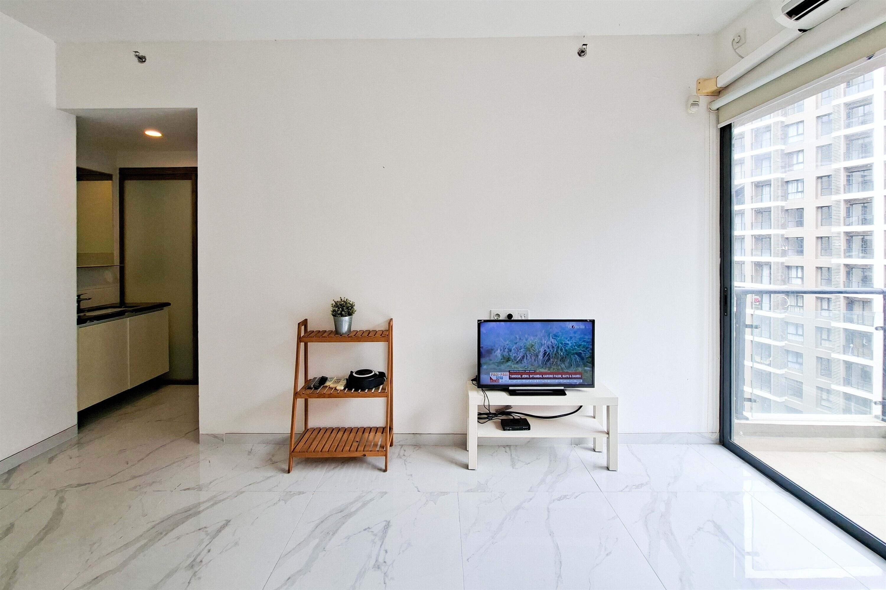 Un salon moderne avec un téléviseur posé sur un meuble, une étagère en bois et une grande fenêtre donnant sur des immeubles.