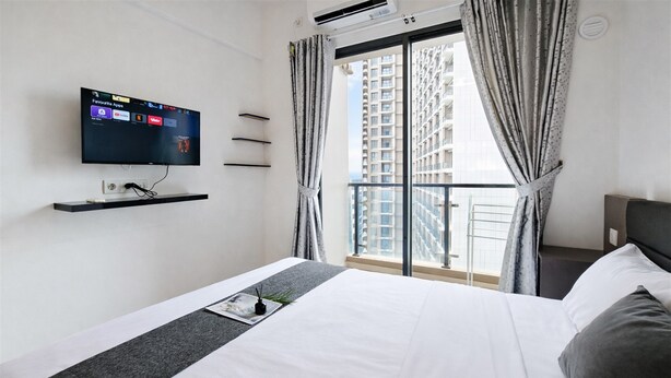 A hotel room with a bed, a TV mounted on the wall, a window with curtains, and a view of tall buildings. - Studio With Simple Style At Sky House Bsd Apartment (Pagedangan)