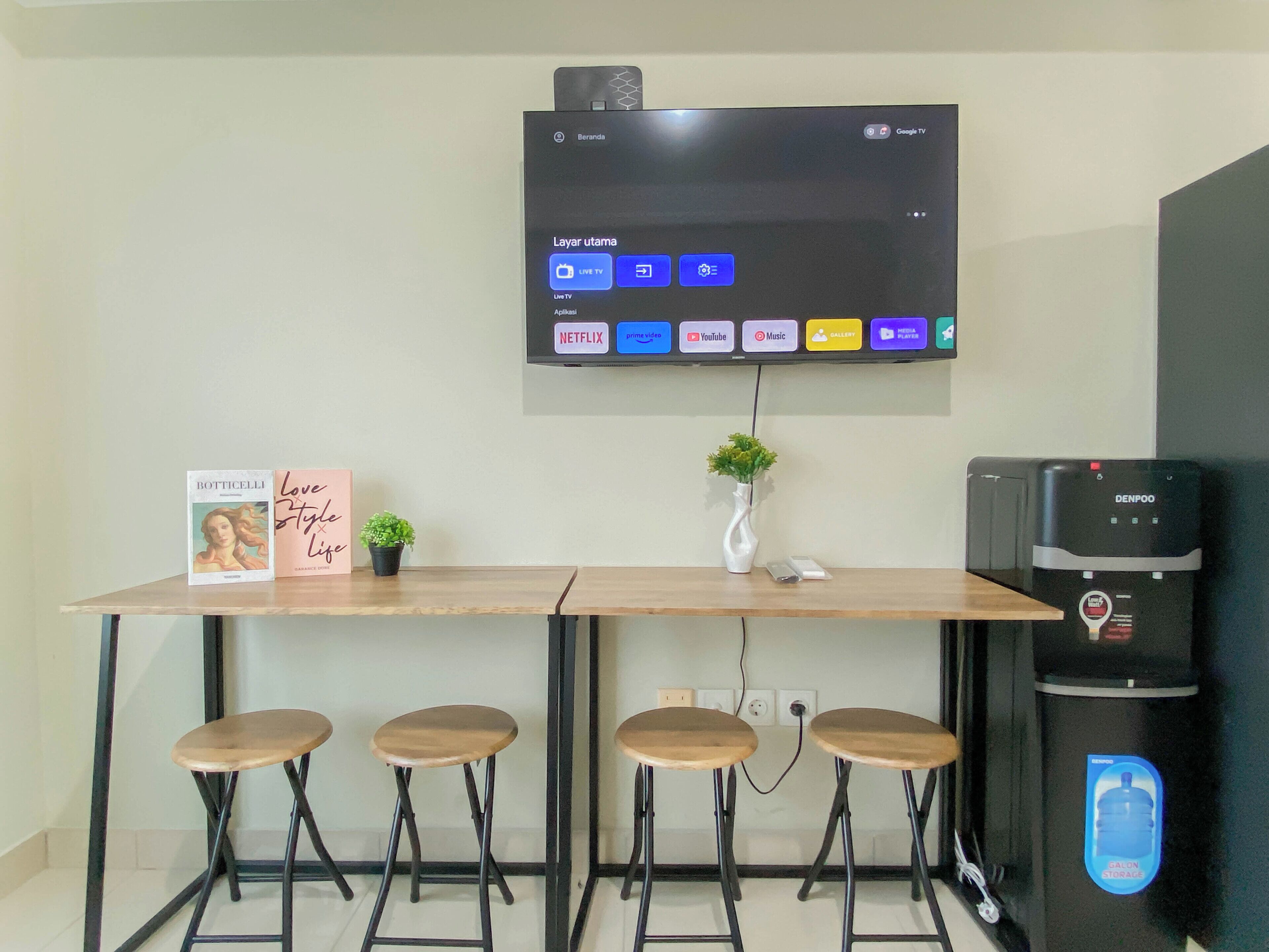 Una cocina moderna con un televisor de pantalla plana sobre una barra de madera, cuatro taburetes, un dispensador de agua y una pequeña planta en maceta.