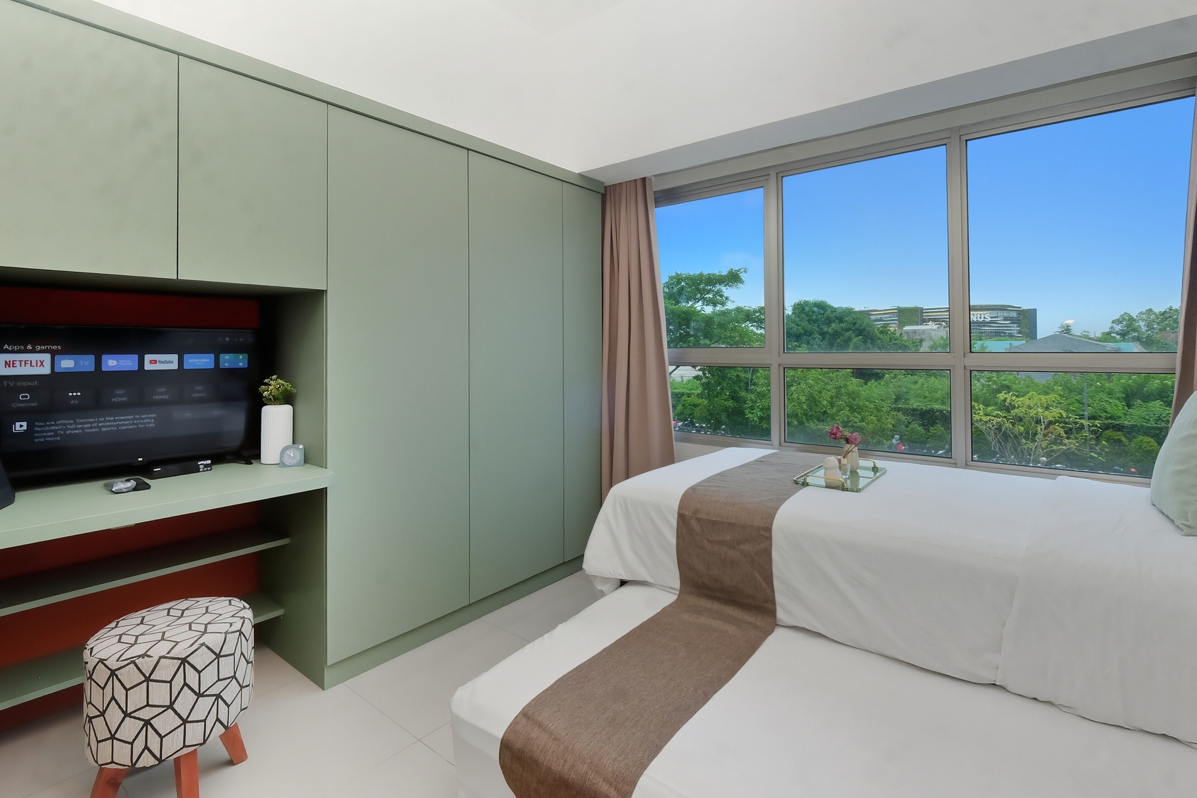 Une chambre d’hôtel moderne, équipée d’un lit, d’un téléviseur posé sur un meuble, d’une fenêtre donnant sur un paysage verdoyant et des bâtiments, ainsi que d’un petit tabouret.