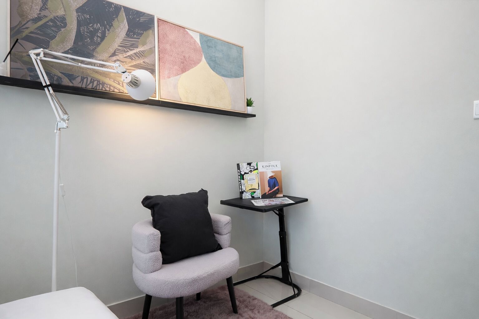 Une petite pièce avec une chaise blanche, un coussin noir, un bureau avec un livre et une lampe sur pied.