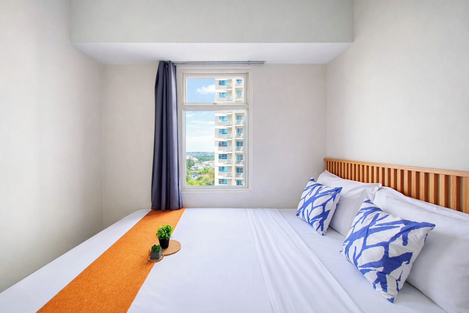 Ein Hotelzimmer mit Bett, Fenster mit Blick auf Gebäude, Holz-Kopfstück und einer kleinen Pflanze auf dem Bett.