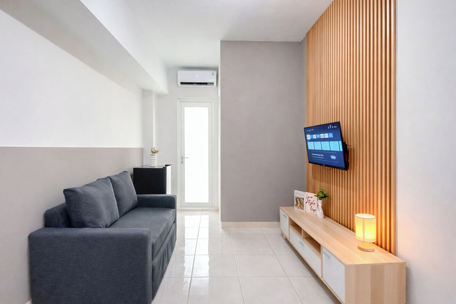 A modern living area with a grey sofa, a wooden panel wall, a mounted TV, and a small table with a lamp.