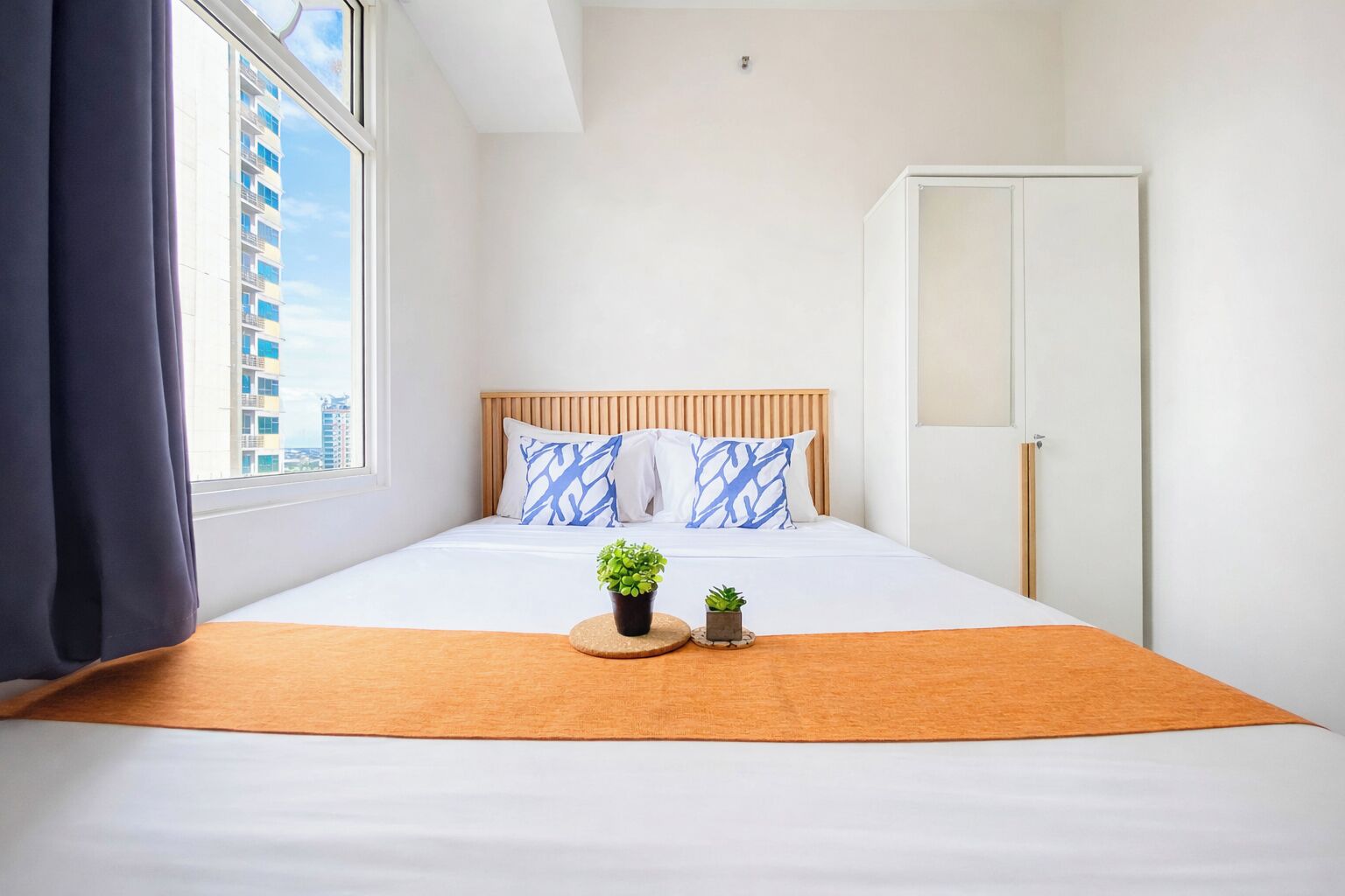 A hotel room with a bed, a window, a door, and a view of buildings outside.
