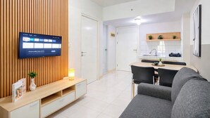 A modern living room with a flat-screen TV, a wooden panel wall, a sofa, a dining area with a table and chairs, and a kitchen area with a sink and shelves. - Fully Furnished 2Br Apartment At Springlake Summarecon Bekasi (Bekasi)
