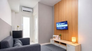 A modern living room with a grey sofa, a TV mounted on a wooden wall, a small wooden side table with a lamp, and a white cabinet. - Fully Furnished 2Br Apartment At Springlake Summarecon Bekasi (Bekasi)
