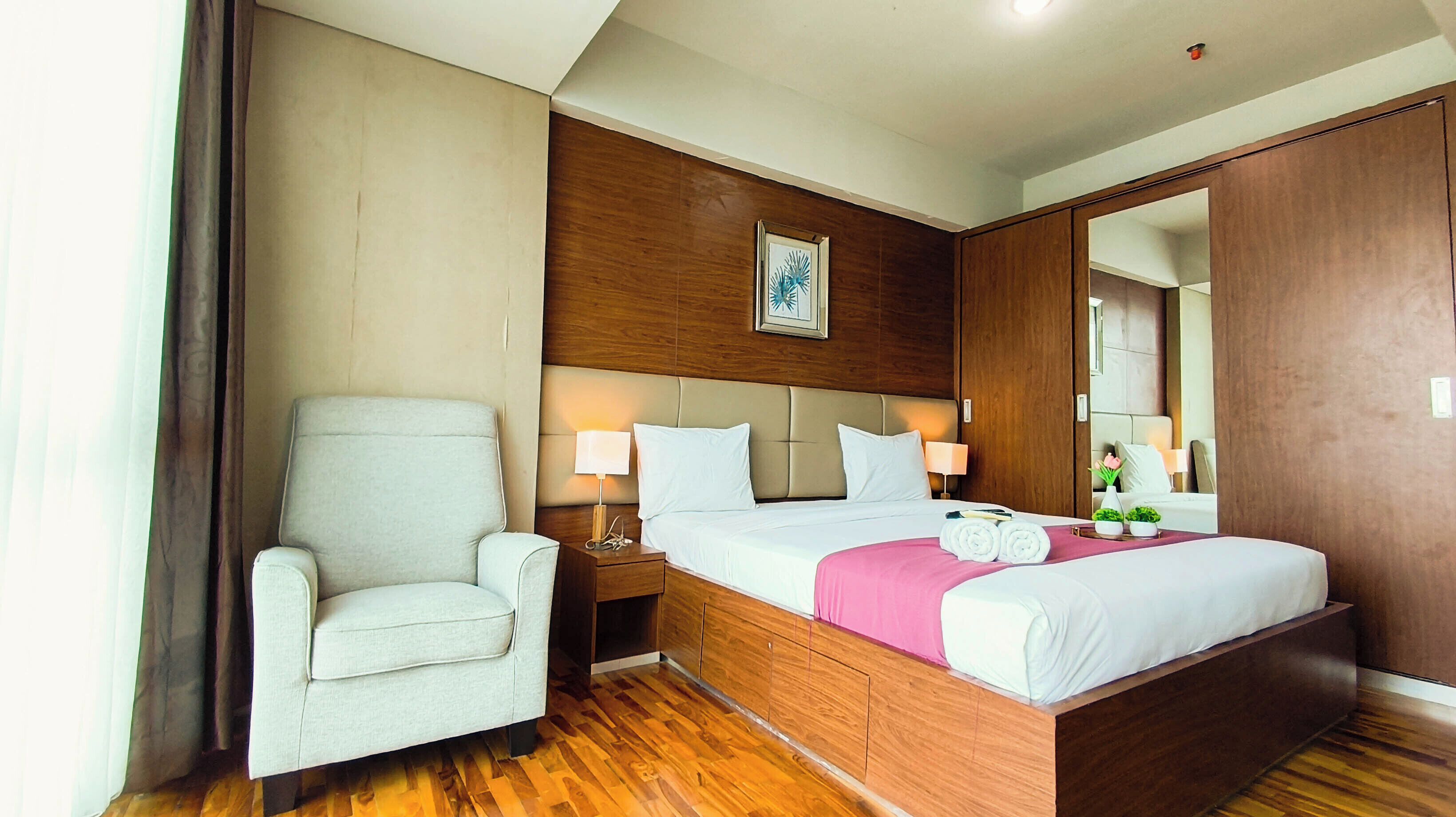 A hotel room with a bed, a chair, a nightstand, a mirror, and a wooden floor.