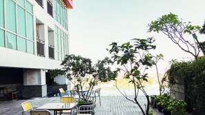 En udendørs terrasse med borde og stole, omgivet af plantekrukker og en bygning med store vinduer.