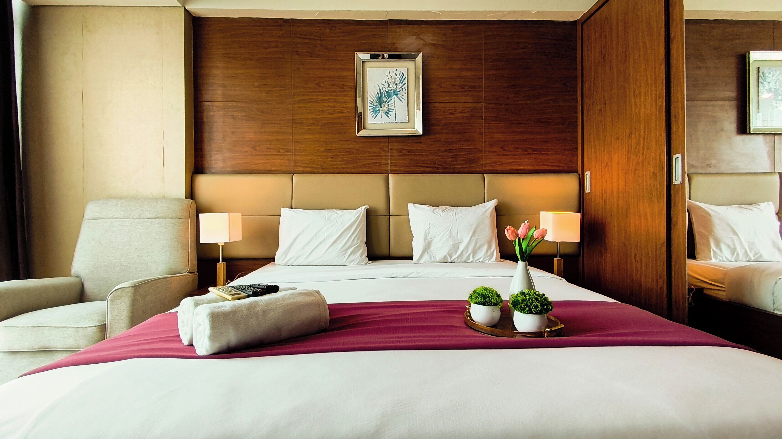 Une chambre d’hôtel avec un grand lit, deux lampes de chevet, une chaise, une petite table sur laquelle est posé un vase de fleurs, et un tableau encadré au mur.