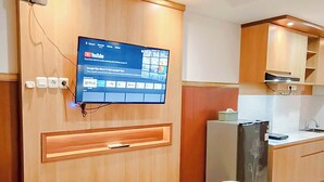 A hotel room with a flat-screen TV mounted on the wall, a bed with a white comforter, a small kitchen area with a refrigerator and a sink, and wooden cabinetry. - Wooden Touch Studio At Tamansari Skylounge Makassar Apartment (Makassar)