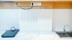 A kitchen with wooden cabinets, a white tiled backsplash, a sink with a faucet, and a power outlet with a cord. - Wooden Touch Studio At Tamansari Skylounge Makassar Apartment (Makassar)