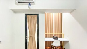 A room with a white desk, a chair, an air conditioning unit, a door with curtains, and a wooden floor. - Modern Look Studio At The Alton Apartment (Semarang)