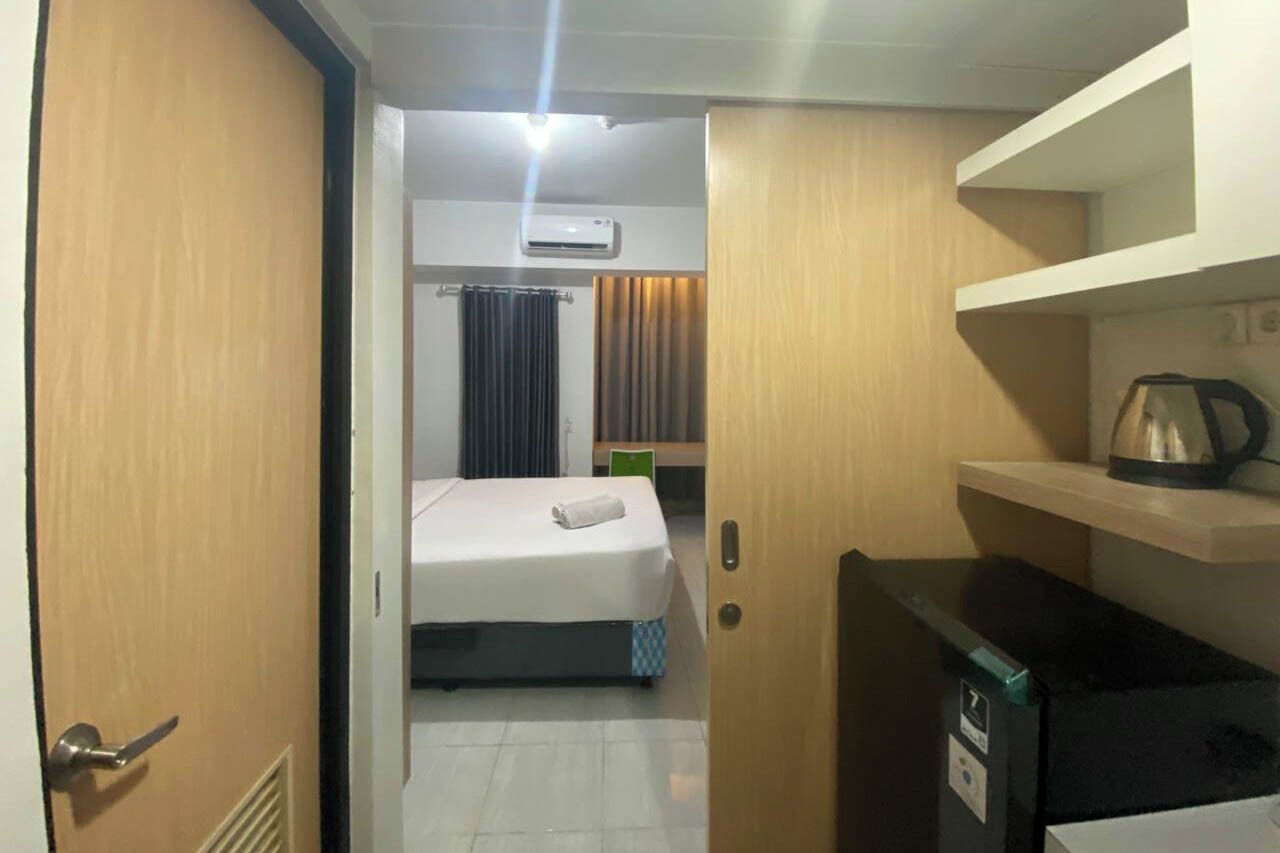 A hotel room with a bed, a small kitchenette, a shelf with a kettle, and an air conditioning unit.